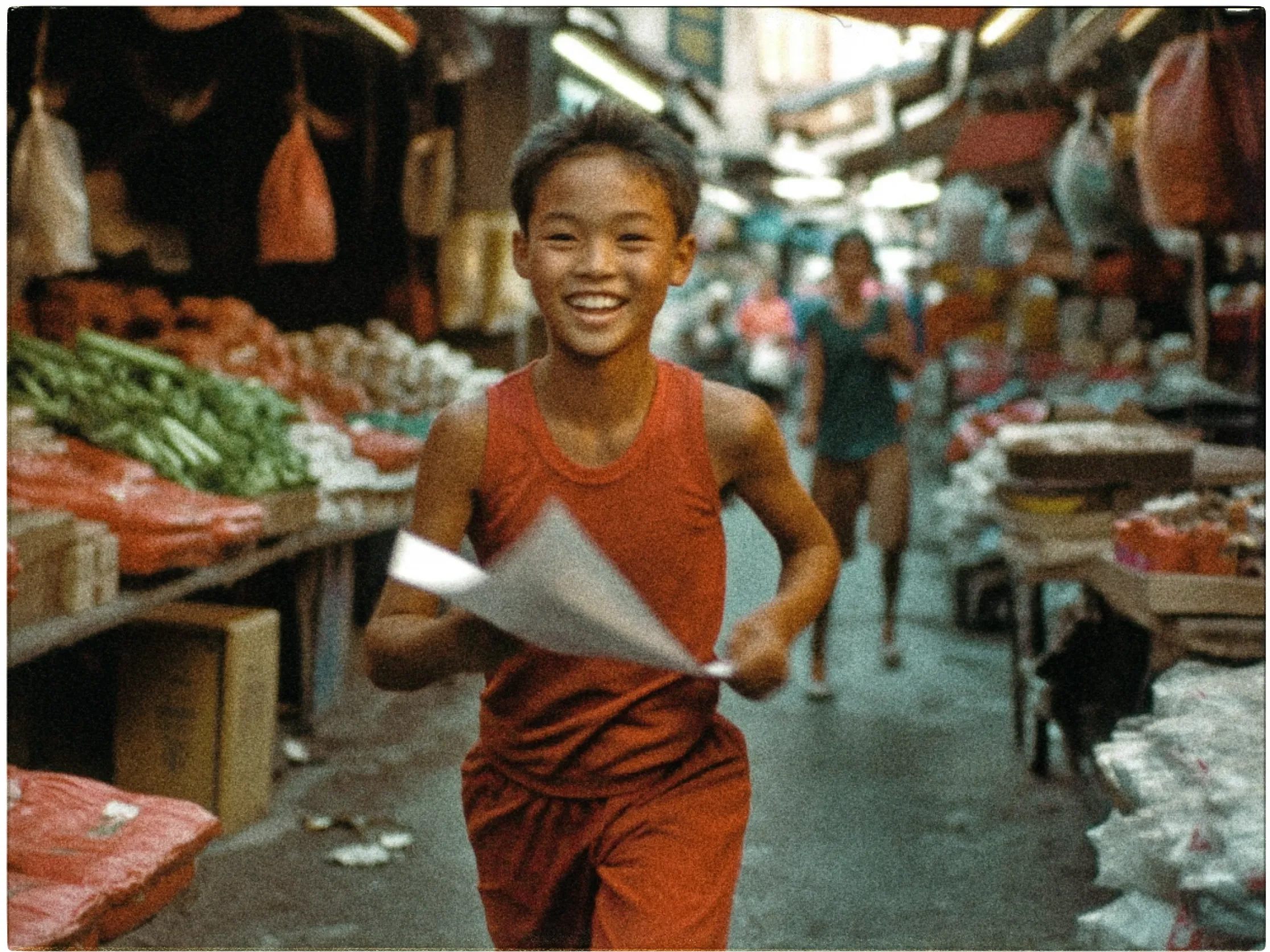 A young boy is captured running joyfully through a bustling market street. He is wearing a sleeveless red shirt and shorts, and in his hand, he holds a sheet of paper. His wide smile conveys happiness and excitement. The market is lined with various stalls displaying fresh produce, including green vegetables and other goods in red wrappings. The background shows blurred figures of people and colorful awnings, contributing to the vibrant, lively atmosphere of the scene. The image has a warm, nostalgic tone, highlighting the dynamic energy of the market.