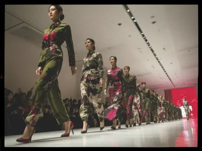 A group of models walks down a brightly lit runway in a fashion show. They wear military-style outfits adorned with vibrant floral patterns in shades of pink and green. The models are lined up neatly and appear focused, with some wearing high-heeled shoes. The audience, visible on the left, watches attentively. The venue has a modern design with a long ceiling light fixture. A red backdrop at the end of the runway enhances the dramatic atmosphere.