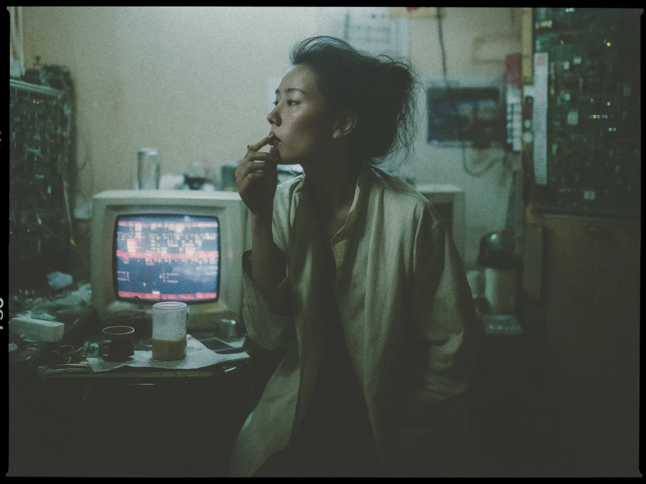 A person sits in a dimly lit room with an old computer monitor in the background displaying unreadable text or code. The individual has short, tousled hair and is wearing a loose, light-colored garment. They are holding an object near their lips, possibly a cigarette, and gazing thoughtfully into the distance. The room is cluttered with various electronic components, wires, and a cup on a table. The overall atmosphere is moody and reflective, with a retro, vintage feel.
