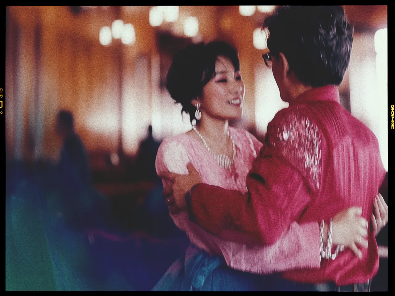 A couple dances closely together in a warmly lit room. The woman, wearing a pink blouse with lace details and pearl jewelry, smiles as she looks at her partner. The man is dressed in a textured red shirt, and they appear to be enjoying a tender moment. The background is softly blurred, with glowing lights creating a romantic ambiance. Their expressions convey joy and connection.