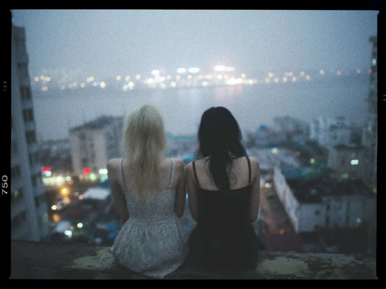 Two people are sitting side by side on a ledge overlooking a cityscape at dusk. The person on the left has light hair and is wearing a light-colored dress, while the person on the right has dark hair and is wearing a black dress. The background features a softly focused city with bright lights reflecting on a body of water, creating a serene and atmospheric scene. The image has a grainy, film-like quality, enhancing the mood.