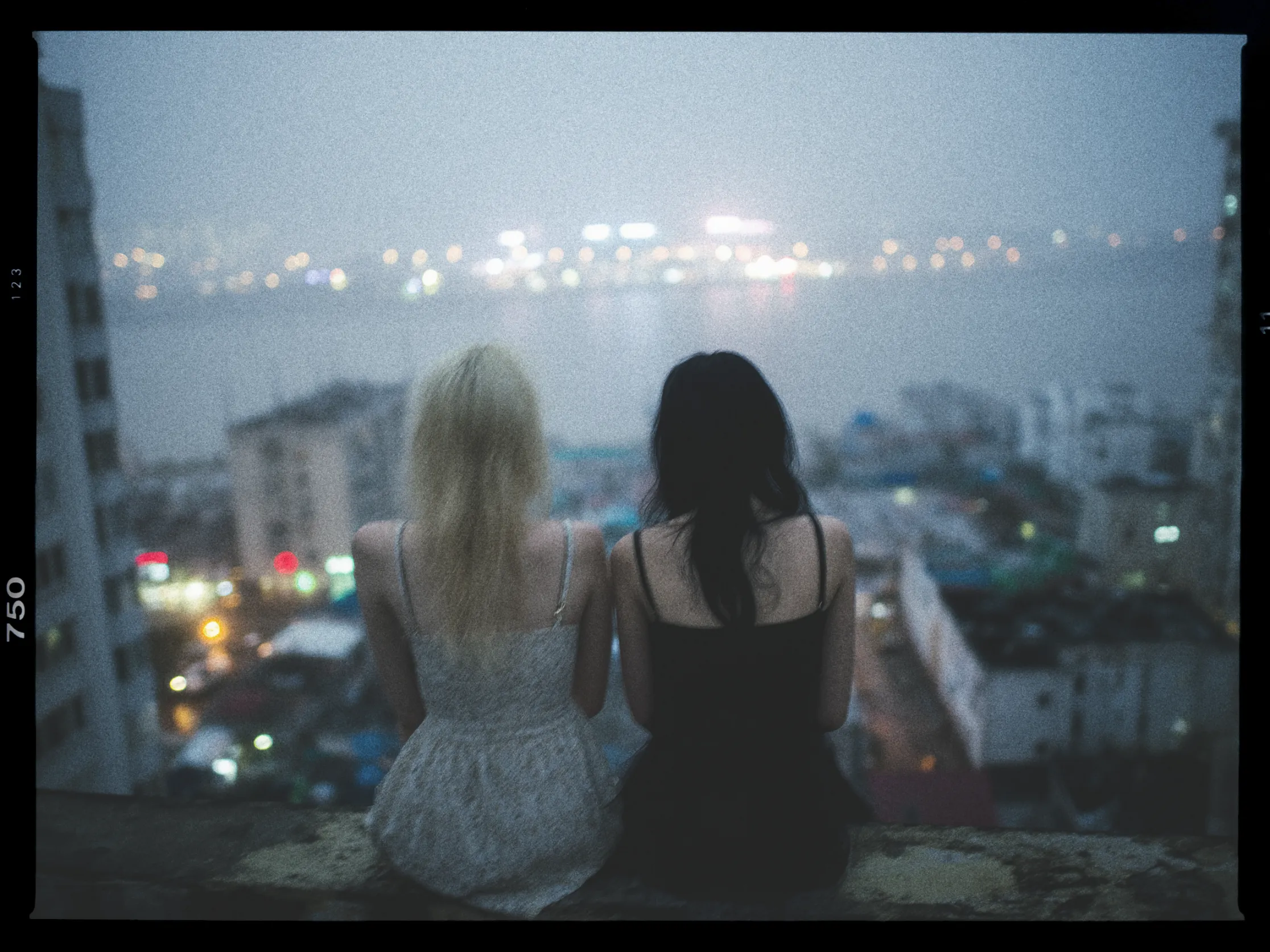 Two people are sitting side by side on a ledge overlooking a cityscape at dusk. The person on the left has light hair and is wearing a light-colored dress, while the person on the right has dark hair and is wearing a black dress. The background features a softly focused city with bright lights reflecting on a body of water, creating a serene and atmospheric scene. The image has a grainy, film-like quality, enhancing the mood.