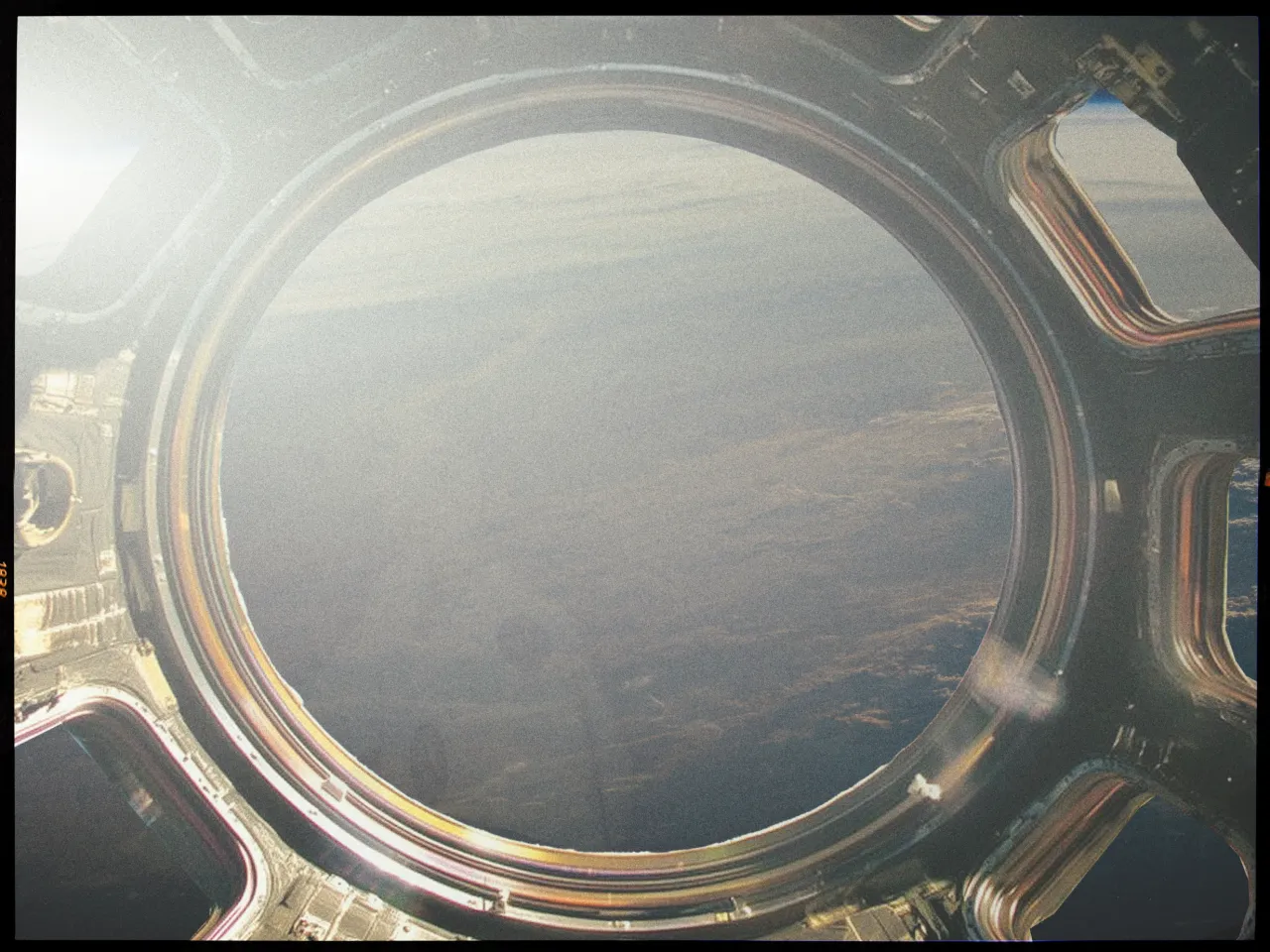A circular window, possibly from a spacecraft, frames a view of Earth from space. The image shows a vast expanse of darkened terrain with a faint, hazy atmosphere and cloud formations visible near the horizon. The metallic edges of the window are highlighted in different colors due to the reflection of light, hinting at the structure's material and design. The image has a grainy quality, suggesting it might have been taken with a film camera or has been digitally altered for artistic effect. The perspective gives the impression of floating in orbit, looking down on the planet.