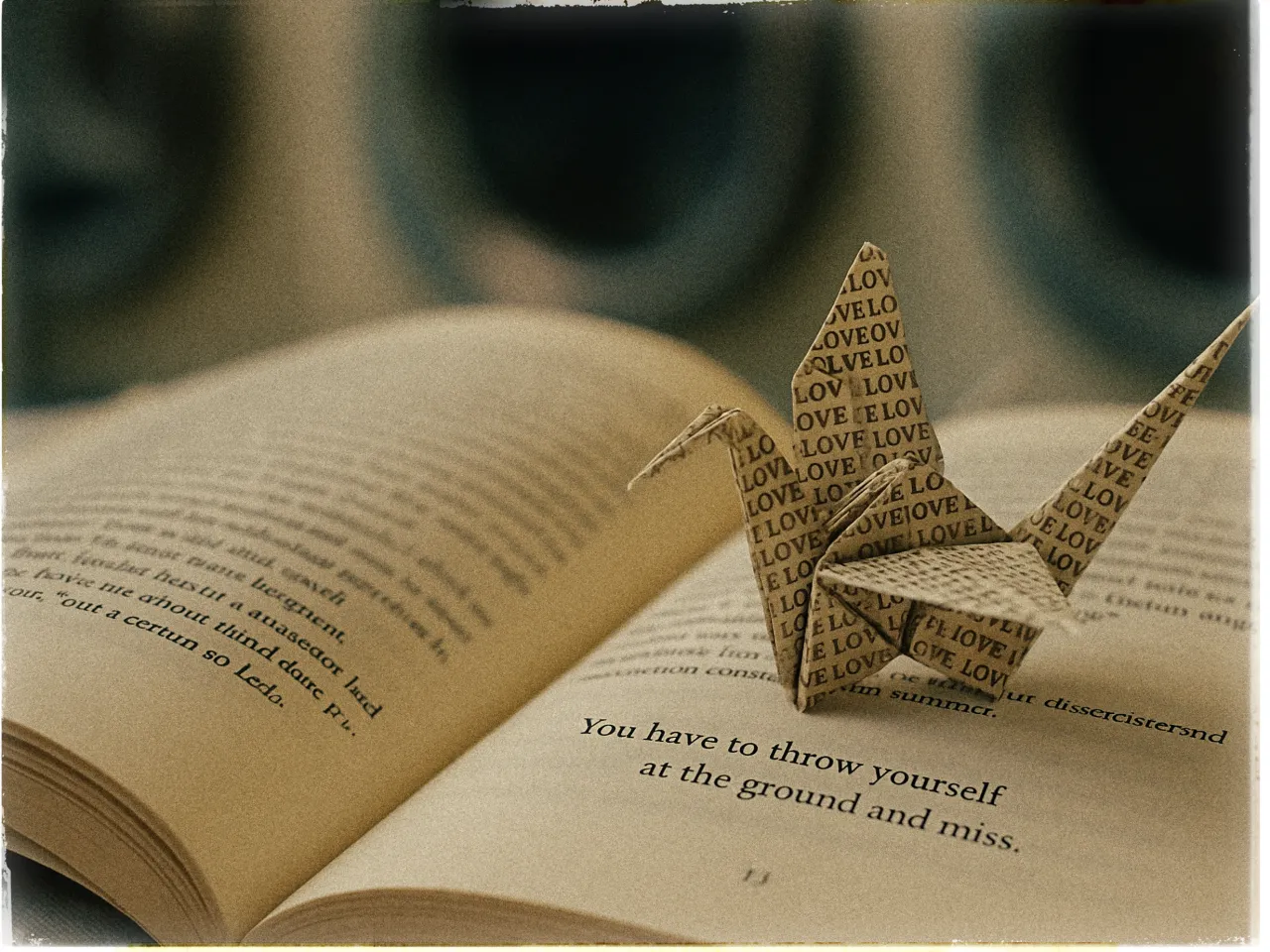 The image shows an open book with pages slightly crumpled. Resting on the pages is an origami crane made from paper printed with the word LOVE repeated in a pattern. The text on the visible page of the book reads, You have to throw yourself at the ground and miss. The background is softly blurred, emphasizing the book and origami in the foreground. The overall tone of the image is warm and nostalgic.