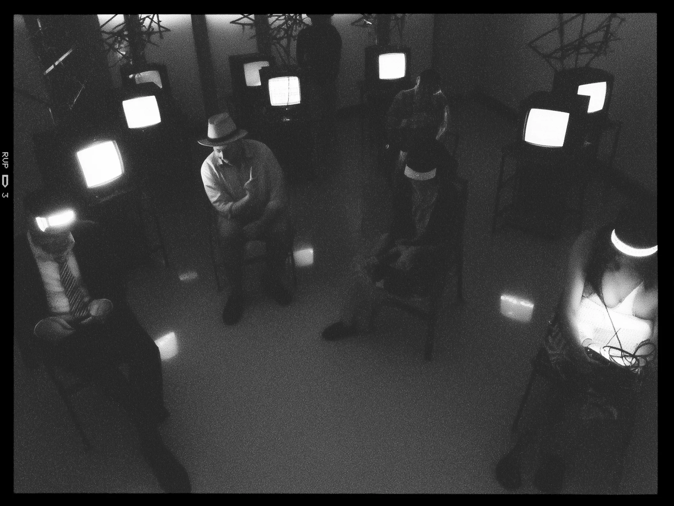 A black and white image depicts a dimly lit room with five people sitting on chairs spaced apart. Each individual is wearing a wide-brimmed hat with a glowing band, casting light onto their faces. They appear to be positioned in a circle, facing inward. Around them, several old-fashioned televisions are placed, some of which have their screens turned on, emitting bright light into the room. The room is minimalistic, with reflective flooring and some abstract metal structures near the walls. Shadows and contrasts are prominent, creating an enigmatic and artistic atmosphere.