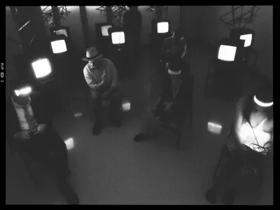 A black and white image depicts a dimly lit room with five people sitting on chairs spaced apart. Each individual is wearing a wide-brimmed hat with a glowing band, casting light onto their faces. They appear to be positioned in a circle, facing inward. Around them, several old-fashioned televisions are placed, some of which have their screens turned on, emitting bright light into the room. The room is minimalistic, with reflective flooring and some abstract metal structures near the walls. Shadows and contrasts are prominent, creating an enigmatic and artistic atmosphere.