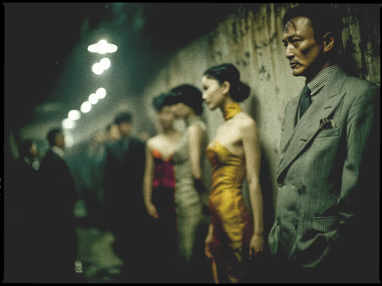 In a dimly lit, atmospheric scene, a group of elegantly dressed individuals stand against a textured wall. In the foreground, a man in a sharp gray pinstripe suit with a pocket square looks pensive, with his hands in his pockets. Beside him, a woman in a striking gold strapless dress stands with her hair styled up. The background is filled with more people, slightly out of focus, wearing formal attire. Overhead lights create a moody ambiance as they recede into the distance, casting a subtle glow on the scene.