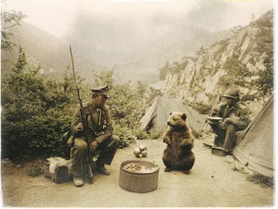 A sepia-toned photograph shows two soldiers in military uniforms sitting in a makeshift camp surrounded by mountains. The soldier on the left is holding a rifle and looking at a bear cub sitting on its haunches in front of a large tin container filled with food. The soldier on the right is crouched by a tent, holding a bowl. There is foliage and rocky terrain in the background, suggesting a remote, mountainous location. The bear appears calm and is the central figure among the humans.