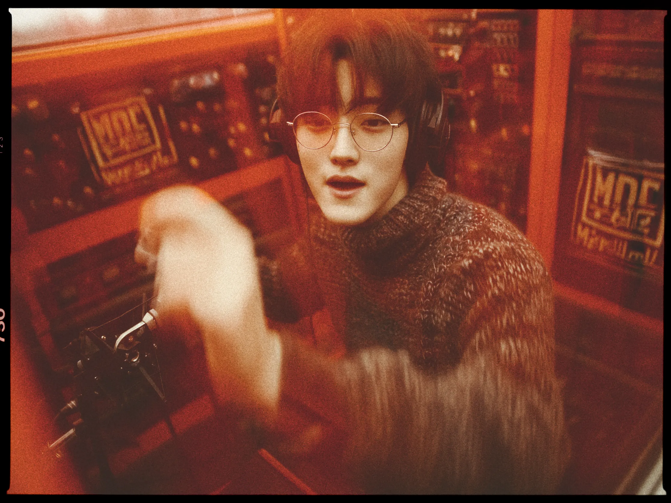 A person is inside a soundproof booth, wearing large headphones and round glasses. They have a short, dark hairstyle and are wearing a thick, textured sweater. The person appears to be engaged in a recording session, with one hand raised in motion, possibly gesturing while speaking or singing into a microphone placed in front of them. The booth is filled with sound equipment, and the lighting has a warm, reddish hue, giving the image a vintage feel. The expression on their face suggests focus and engagement.