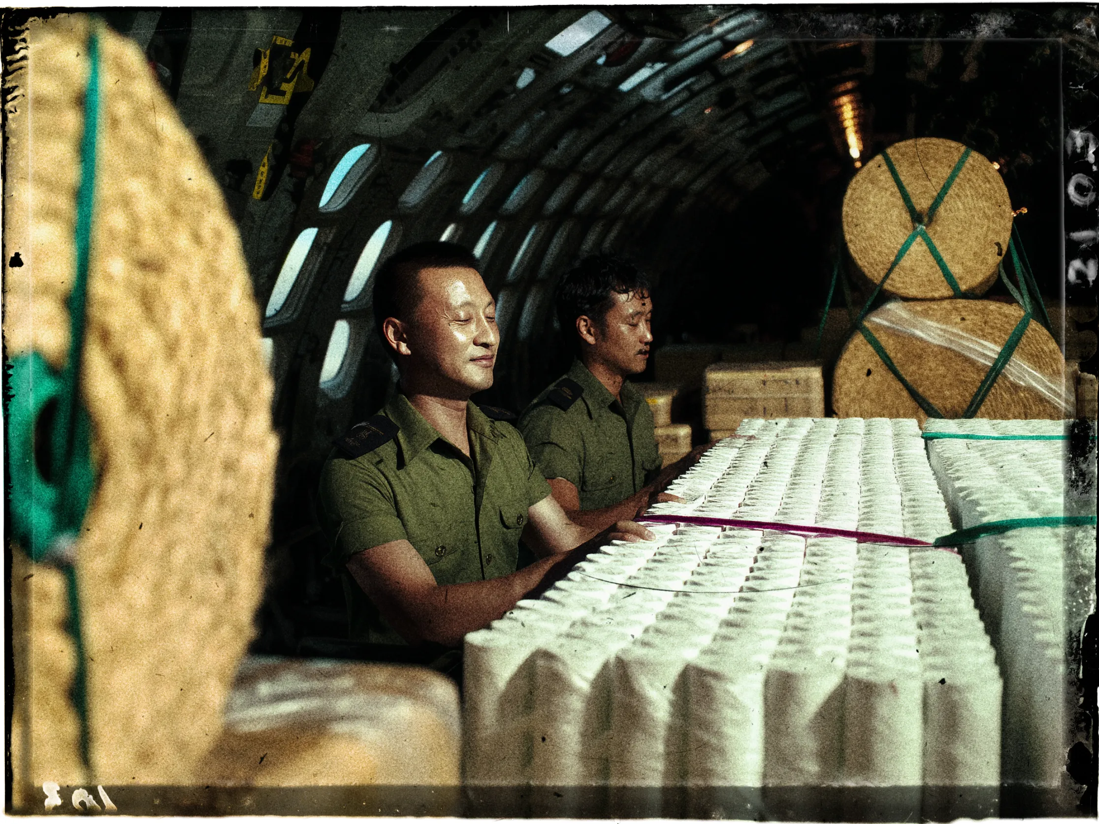 Two individuals in uniform are sitting inside an aircraft's cargo area, surrounded by large bales and stacks of packages. The interior of the aircraft is lined with evenly spaced windows, casting a pattern of light. In the foreground, rows of tightly packed white cylindrical items are strapped together with colored bands. The lighting is mixed, highlighting the individuals and casting shadows, creating a somber atmosphere.