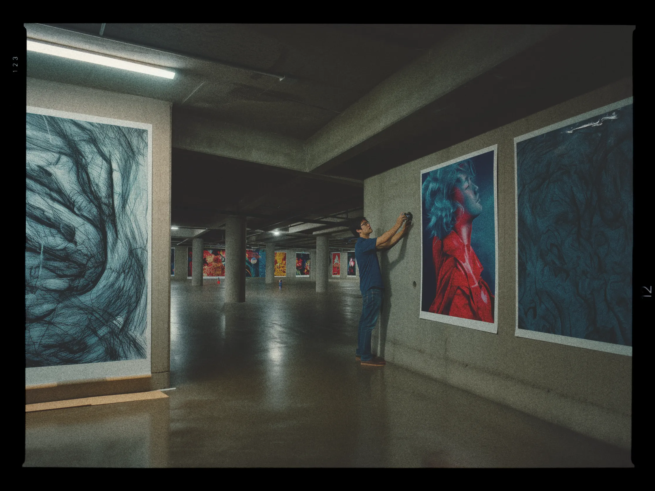A person is positioned in an expansive, dimly lit gallery space where they are hanging large, vibrant art pieces on concrete walls. The art includes abstract designs and a striking portrait of a person with their head tilted back, sporting white hair and a red coat. The gallery features a series of similarly bold and colorful artworks along the walls in the background, illuminated by overhead lights. The floor is a polished concrete, reflecting the muted lighting. The scene conveys a sense of preparation for an exhibition.