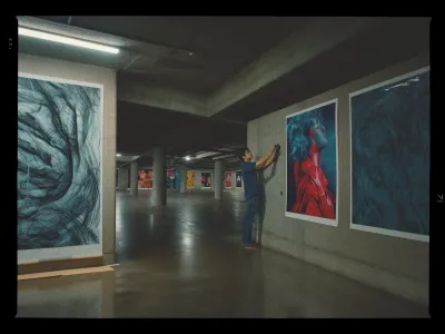 A person is positioned in an expansive, dimly lit gallery space where they are hanging large, vibrant art pieces on concrete walls. The art includes abstract designs and a striking portrait of a person with their head tilted back, sporting white hair and a red coat. The gallery features a series of similarly bold and colorful artworks along the walls in the background, illuminated by overhead lights. The floor is a polished concrete, reflecting the muted lighting. The scene conveys a sense of preparation for an exhibition.