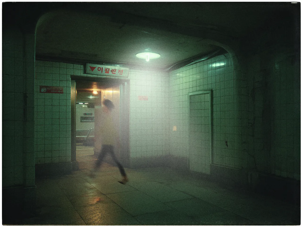 A dimly lit, tiled underpass with a moody atmosphere. The walls and ceiling are covered with white square tiles, some showing signs of wear. A person wearing dark pants and a light shirt is captured in motion, appearing blurred as they walk through the passageway. A glowing, circular light fixture hangs from the ceiling. In the background, a doorway leads to another section, with signage in a foreign script above it. The overall scene is enveloped in a greenish hue, enhancing the mysterious and somewhat eerie feeling.