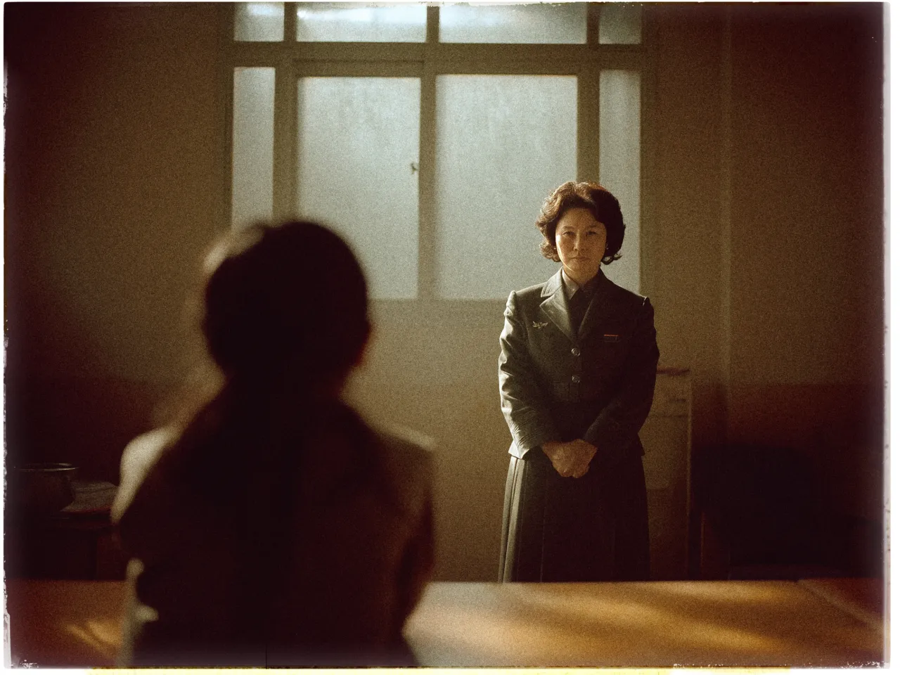The image depicts a dimly lit room with a serious atmosphere. A woman in a formal gray uniform stands near the window with frosted glass, her hands clasped in front of her. Her expression is calm yet authoritative. The room's lighting casts a warm, muted glow, creating a soft, vintage effect. In the foreground, a blurred figure, likely a child or young person, is seated and facing the woman, with their back toward the viewer, suggesting an interaction or confrontation between the two.