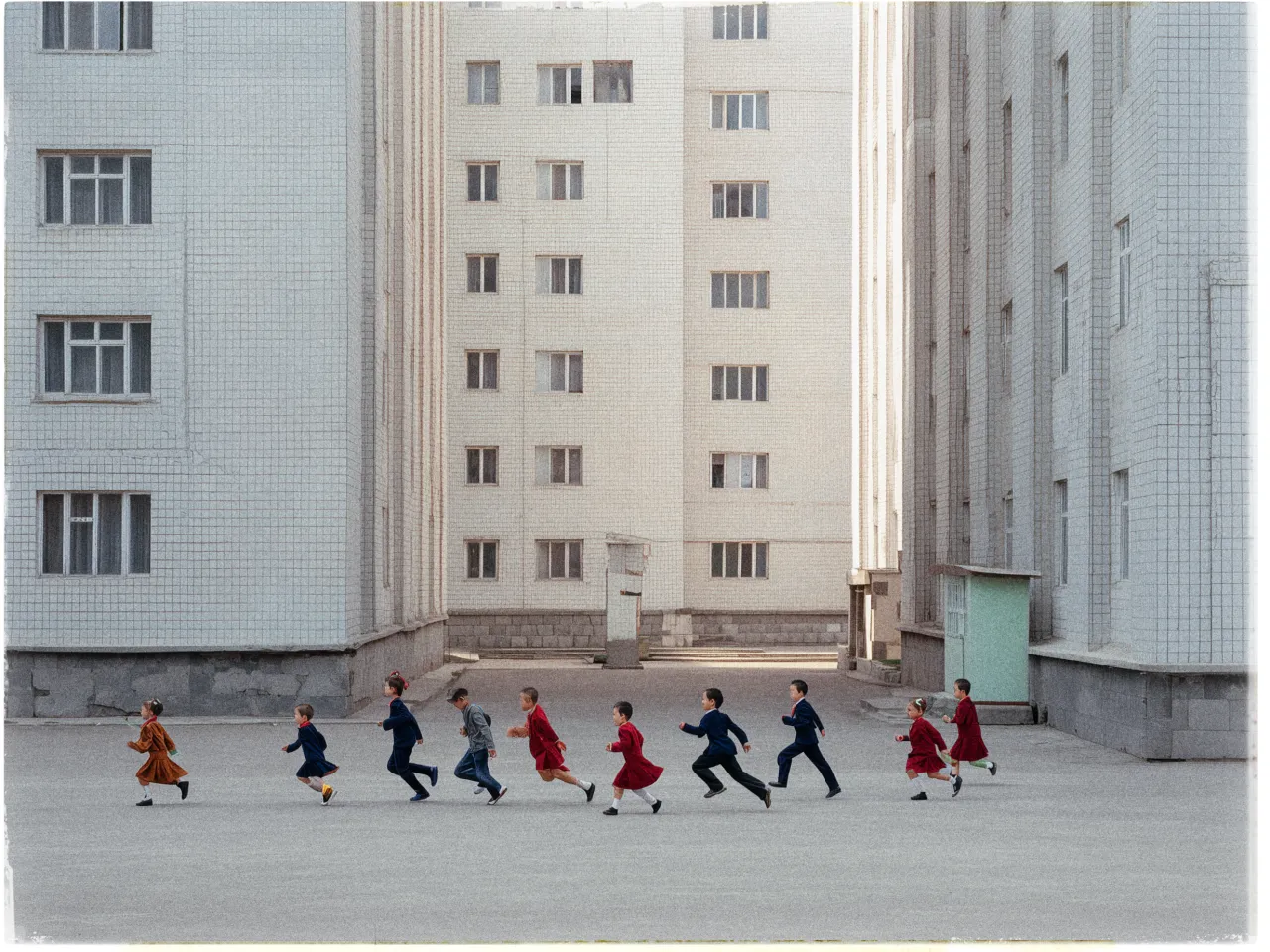 A group of children runs in a line across a gray, empty courtyard surrounded by tall, pale, tiled apartment buildings. The buildings have multiple windows, evenly spaced, imparting a Soviet-era architectural feel. The children, dressed in a mix of vibrant reds and blues, create a lively contrast against the stark, muted background. The courtyard is barren, with the focus solely on the children's movement. The scene conveys a sense of playful energy amid a rigid, urban environment.