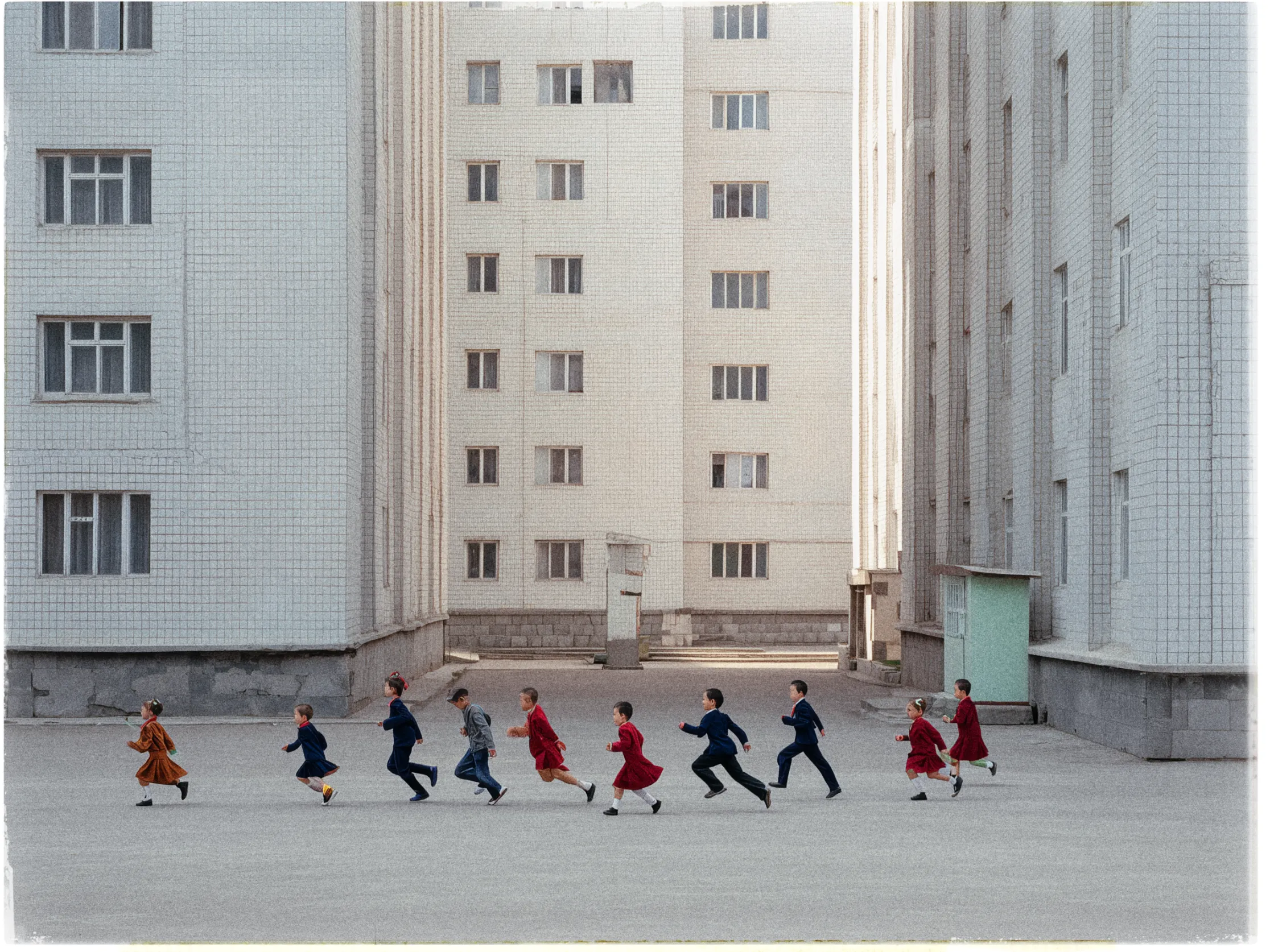 A group of children runs in a line across a gray, empty courtyard surrounded by tall, pale, tiled apartment buildings. The buildings have multiple windows, evenly spaced, imparting a Soviet-era architectural feel. The children, dressed in a mix of vibrant reds and blues, create a lively contrast against the stark, muted background. The courtyard is barren, with the focus solely on the children's movement. The scene conveys a sense of playful energy amid a rigid, urban environment.