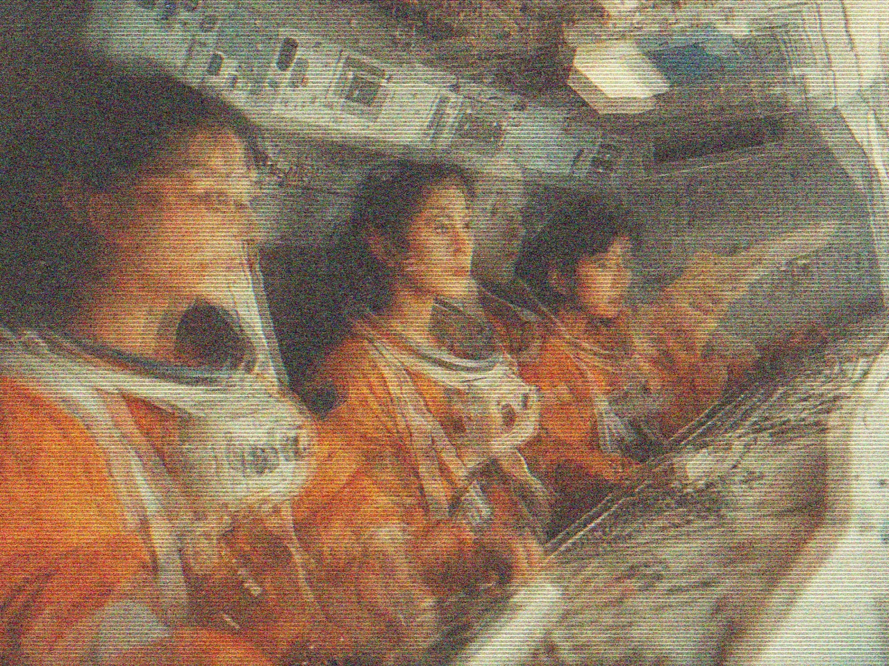 The image shows three people in an astronaut cockpit, wearing orange spacesuits with white helmets resting on their laps. They are seated side by side, facing a complex array of control panels filled with numerous buttons and screens. The image has a blurred effect, giving a sense of motion or multiple exposures. The individuals appear focused, gazing toward the screens in front of them. The interior of the cockpit is metallic and densely packed with technical equipment, creating an authentic spaceflight setting.