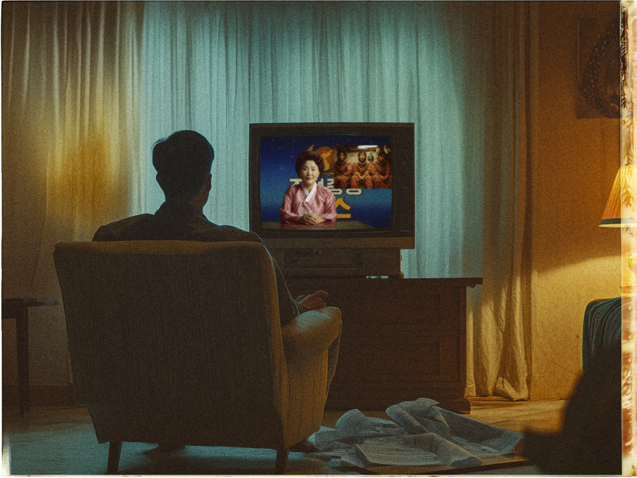 A person is sitting in an armchair in a dimly lit room, facing an old-fashioned television. The TV shows a person in a pink outfit against a blue background with text and images. The room is warmly lit with yellow light from a lamp on the right. Behind the chair, sheer curtains are drawn across a window, filtering the exterior light. Papers or sheets are scattered on the floor in front of the chair, adding a casual, perhaps cluttered feel to the scene.