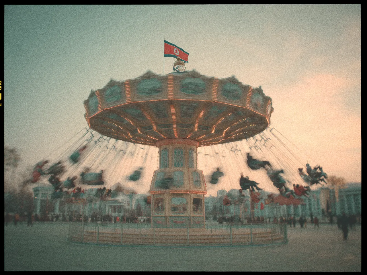 A vintage-filtered image depicts a spinning amusement park swing ride, captured in motion. The central pillar of the ride is ornately decorated, and swings are suspended from chains around the top edge, with riders appearing as blurred figures due to the motion. A flag is visible on top of the structure. The background shows a crowd of people and park structures, under a soft, dusky sky. The overall tone is nostalgic with a grainy texture.