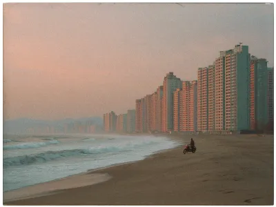 A serene coastal scene at dusk with a wide sandy beach stretching along the left side. The ocean waves gently roll onto the shore, creating a soft, misty foam. On the right, a long row of tall, uniform apartment buildings extends into the distance, bathed in a warm, pinkish hue from the setting sun. A person sits on a motorcycle near the water's edge, adding a sense of solitude and reflection. The distant mountains and a pastel-colored sky enhance the tranquil ambiance.