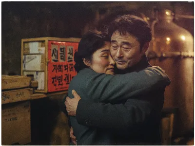 A middle-aged couple is embracing warmly in a dimly lit room. The woman, with closed eyes, rests her head against the man's shoulder, while he gently holds her, exuding a sense of comfort and care. They wear dark coats and appear content. In the background, there are stacked boxes with Korean writing on them, and a large copper-colored distillation apparatus, suggesting an industrial or workshop setting. The lighting casts a cozy, intimate atmosphere over the scene.