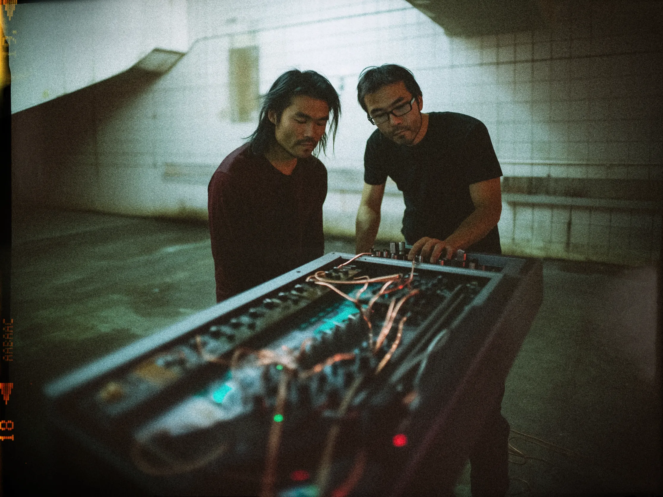 The image features two people focused on a large, complex audio equipment setup in a dimly lit, industrial-like space with tiled walls. The person on the left has long hair and is wearing a dark sweater, while the person on the right, with short hair and glasses, is wearing a black t-shirt. They are both leaning over the equipment, which is covered in numerous wires and knobs, likely indicating a sound mixing or electronic music device. The ambient lighting casts a moody, almost cinematic atmosphere, with a greenish tint and shadows on the wall.