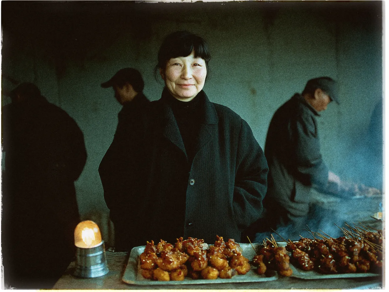 A woman stands behind a street food stall, smiling gently at the camera. She wears a dark coat and has short, dark hair. In front of her are trays filled with skewered, glazed meat, possibly chicken, sprinkled with sesame seeds. The background features a dimly lit environment with two other people; one appears to be a man, tending to a grill, shrouded in subtle smoke. The overall atmosphere is warm and inviting, capturing a candid moment at a bustling street market.