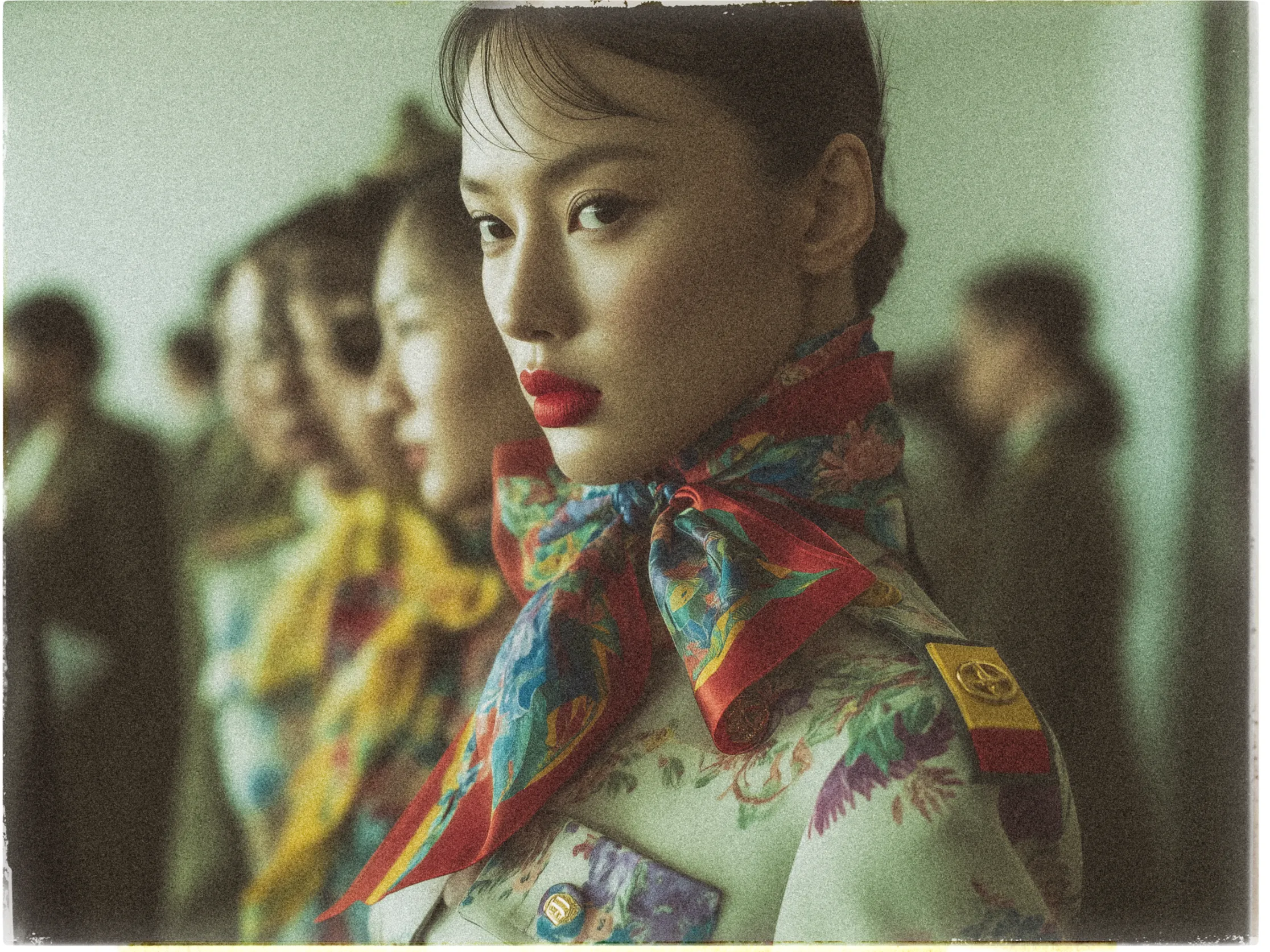 The image features a group of people standing in formation, with a focus on a woman in the foreground. She has striking red lipstick and intense eyes, looking directly at the camera. Her attire is floral-patterned, and she wears a colorful scarf tied around her neck with an elaborate knot. Her outfit includes insignia on the lapel and shoulder, which suggests a uniform. The background is softly blurred, showing other similarly dressed individuals. The overall tone of the image is vintage, characterized by soft, muted colors and a slightly grainy texture.