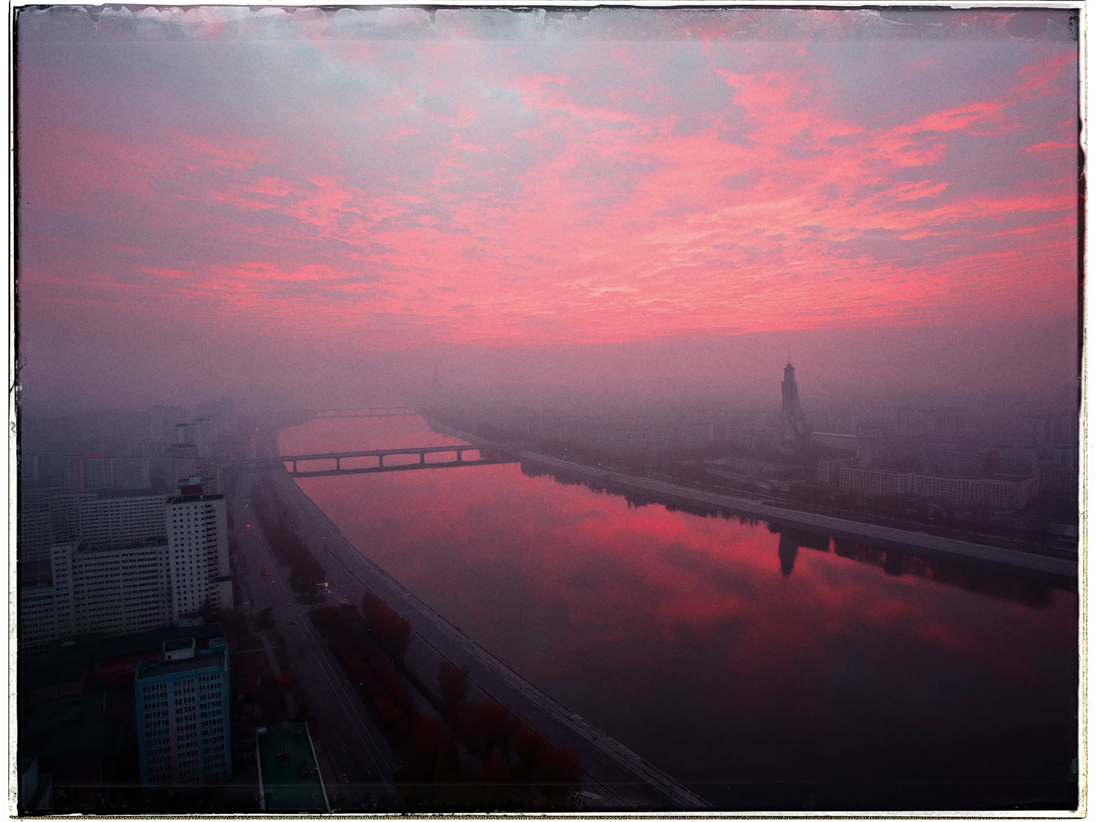 Aerial view of a cityscape at dusk with a striking pink and red sunset sky. The clouds are vividly colored, reflecting off the river that runs through the city. A bridge spans the river, and there are large, silhouetted buildings on both sides, including a tower-like structure on the right. The streets and riverbanks appear quiet and partially obscured by mist or haze, adding a serene and moody atmosphere to the scene.