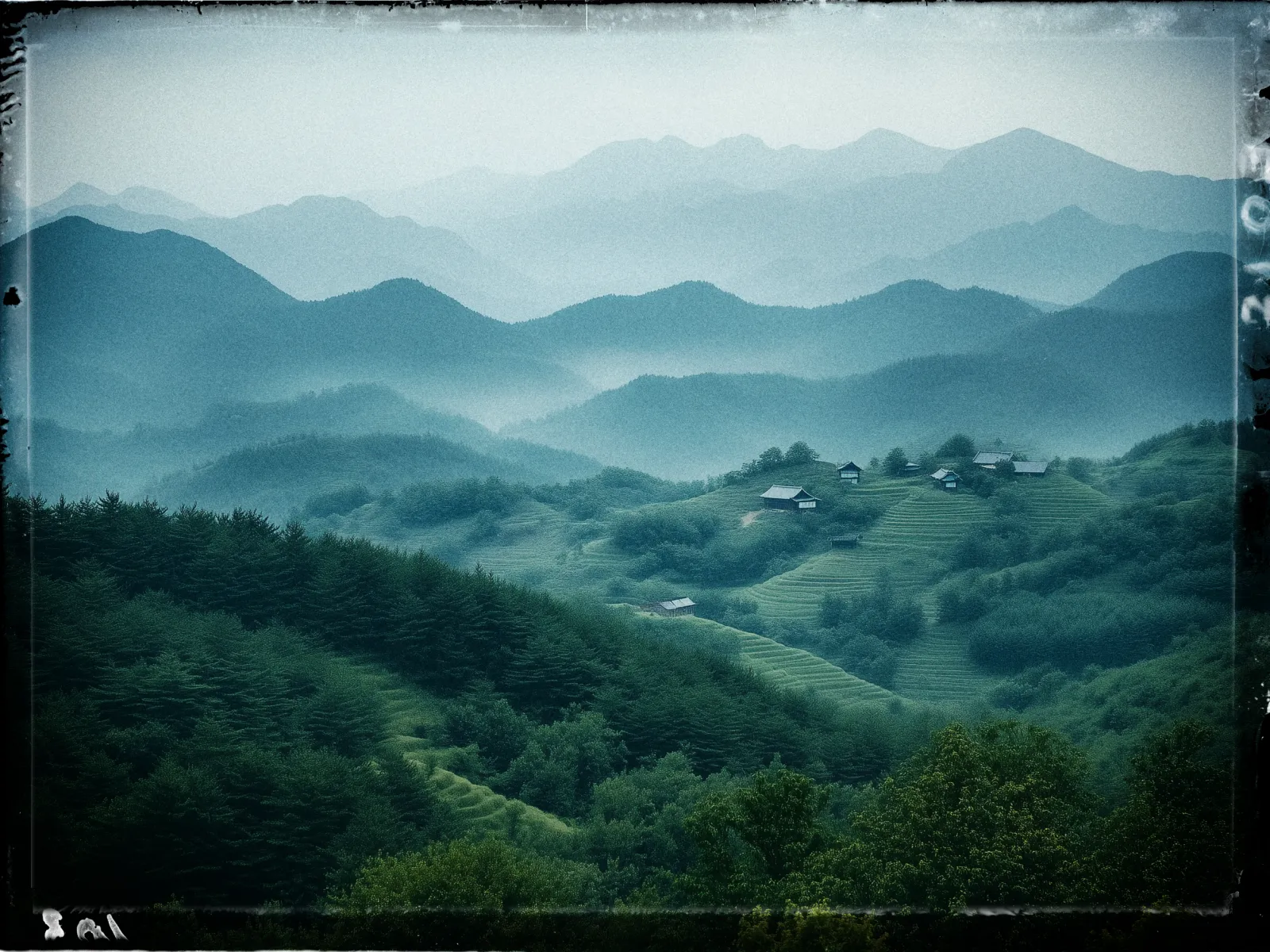 A serene landscape depicts rolling, lush green hills blanketed in dense forests with terraces gently carved into the slopes. Scattered among the greenery are a few small wooden houses, adding a sense of rustic tranquility. In the background, a series of misty, blue-tinted mountain ranges fade gradually into the distance, creating a layered effect. The overall atmosphere is calm and peaceful, with a slightly vintage or film-like quality, enhanced by a subtle border that frames the image.