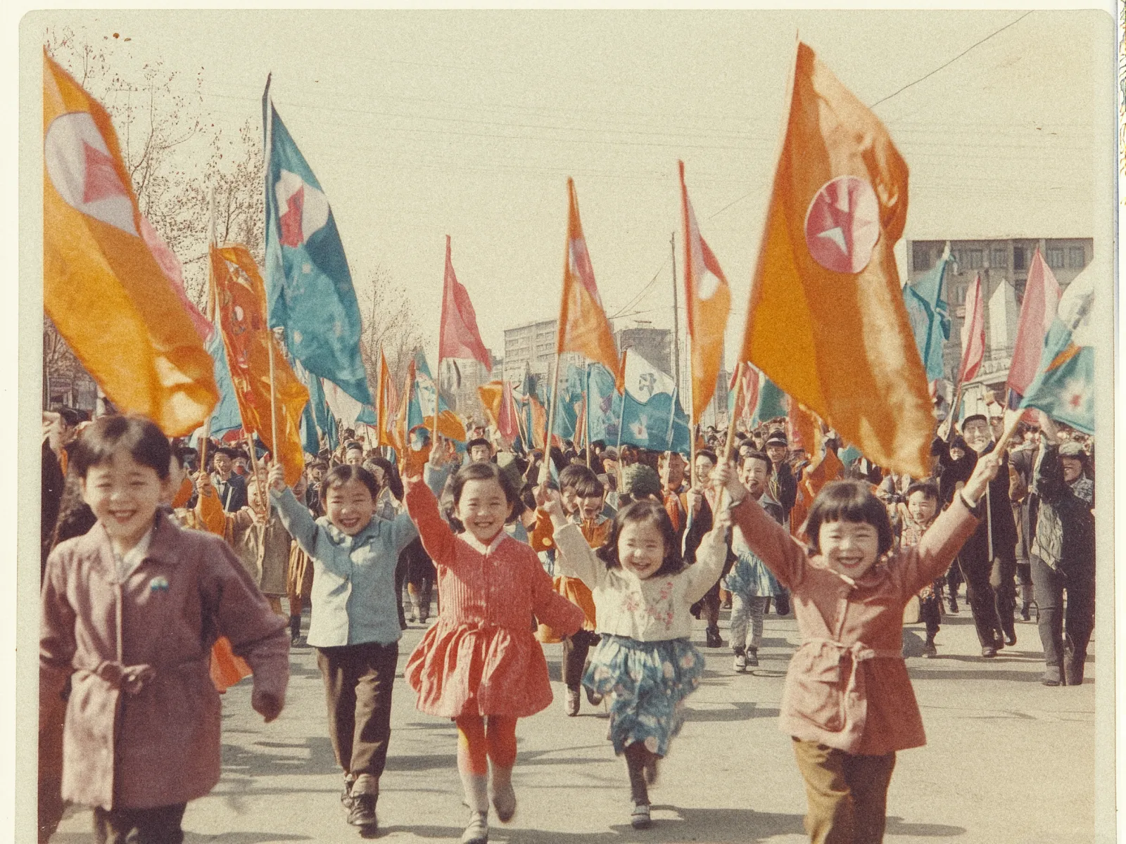 A group of children, smiling and joyous, run toward the camera down a wide street. They are waving vibrant orange and blue flags adorned with a red star emblem. The children are dressed in warm clothing, with some wearing coats and dresses. Behind them, more people follow with similar flags. The scene appears to be in a city, given the presence of multi-story buildings in the background. It's a bright day, and the atmosphere is festive and lively. Bare trees line the street, suggesting it might be autumn or winter.