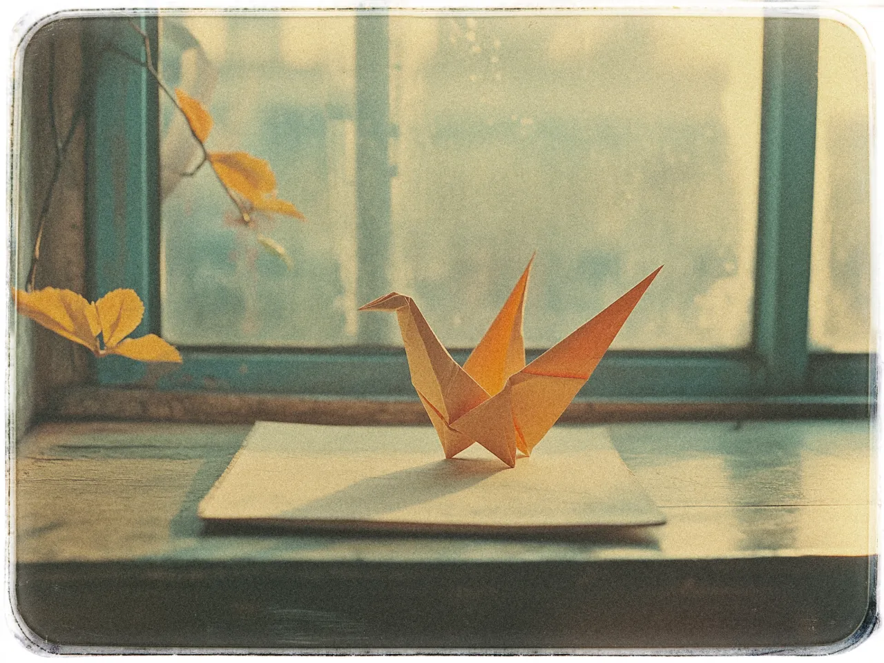 A softly lit image shows an orange origami crane placed on a white piece of paper. The paper rests on a wooden windowsill that has a weathered blue window frame. The window suggests an overcast sky or a gently blurred outdoor view, creating a serene and slightly nostalgic atmosphere. On the left side of the frame, a plant with several yellow leaves is visible, enhancing the warm, autumnal feeling of the scene. The overall tone of the image is warm and calming, with muted colors and soft light.