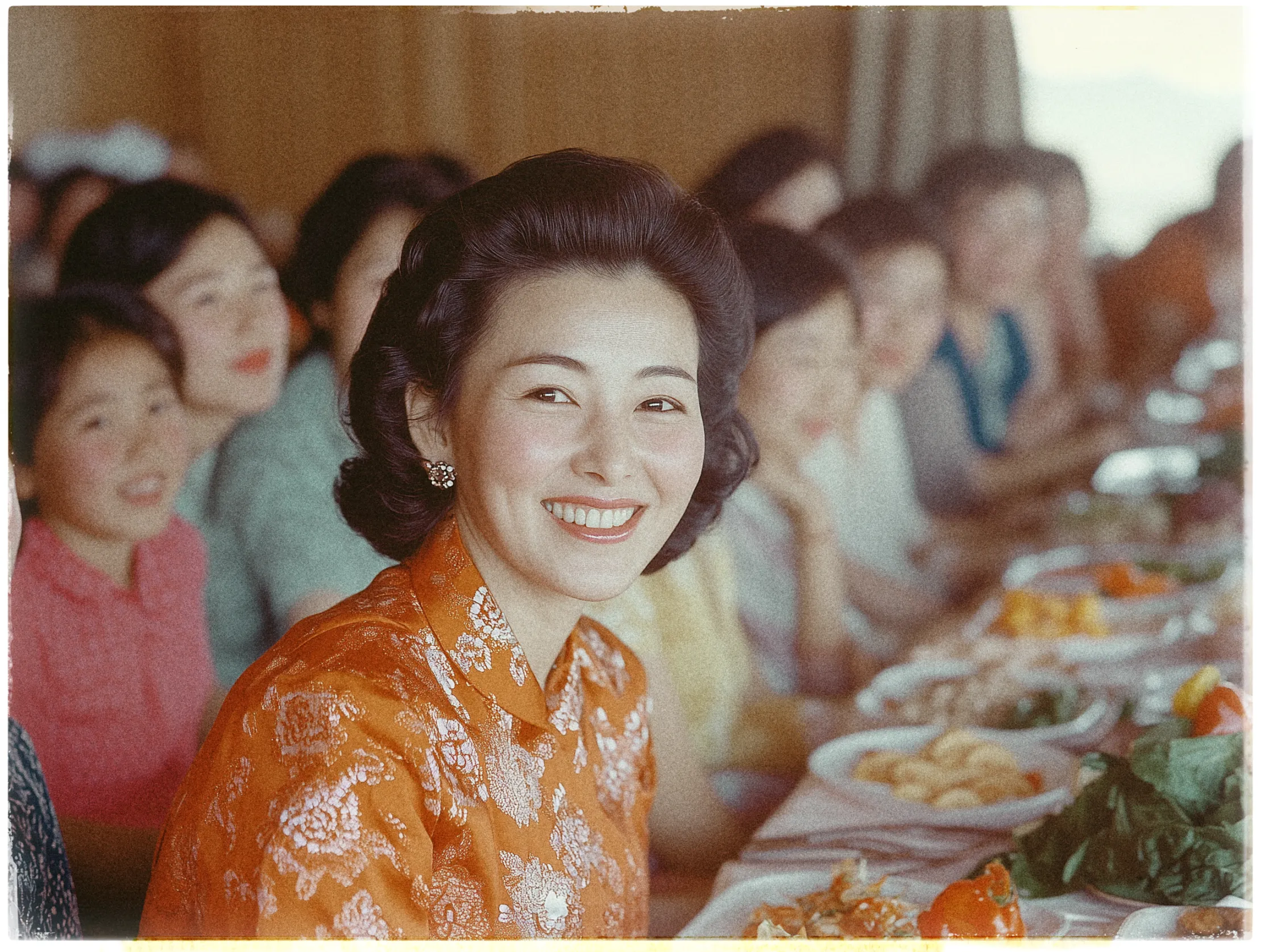 A woman with a warm smile is seated at a dining table, wearing an elegant orange and white floral dress. She has dark, styled hair and is accessorized with pearl earrings. The table before her is laden with various dishes, including plates of vegetables and meats. Other diners, mostly women, are visible along the length of the table, blurred and creating a sense of a lively gathering. The setting appears to be indoors with soft, diffused lighting, suggesting a formal or celebratory occasion.