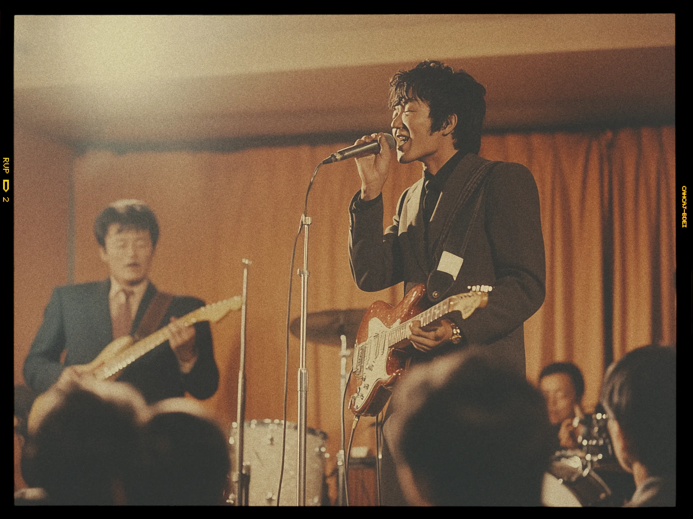 A band performs on a dimly lit stage with an orange curtain backdrop. The focus is on a singer playing an electric guitar, holding a microphone close to his mouth. He wears a dark suit and appears to be passionately singing. To his left, another musician plays a guitar, also wearing a dark suit. A drum set is partially visible in the background. The foreground shows the silhouettes of audience members watching the performance. The overall atmosphere appears vintage, with a warm, sepia-toned filter.