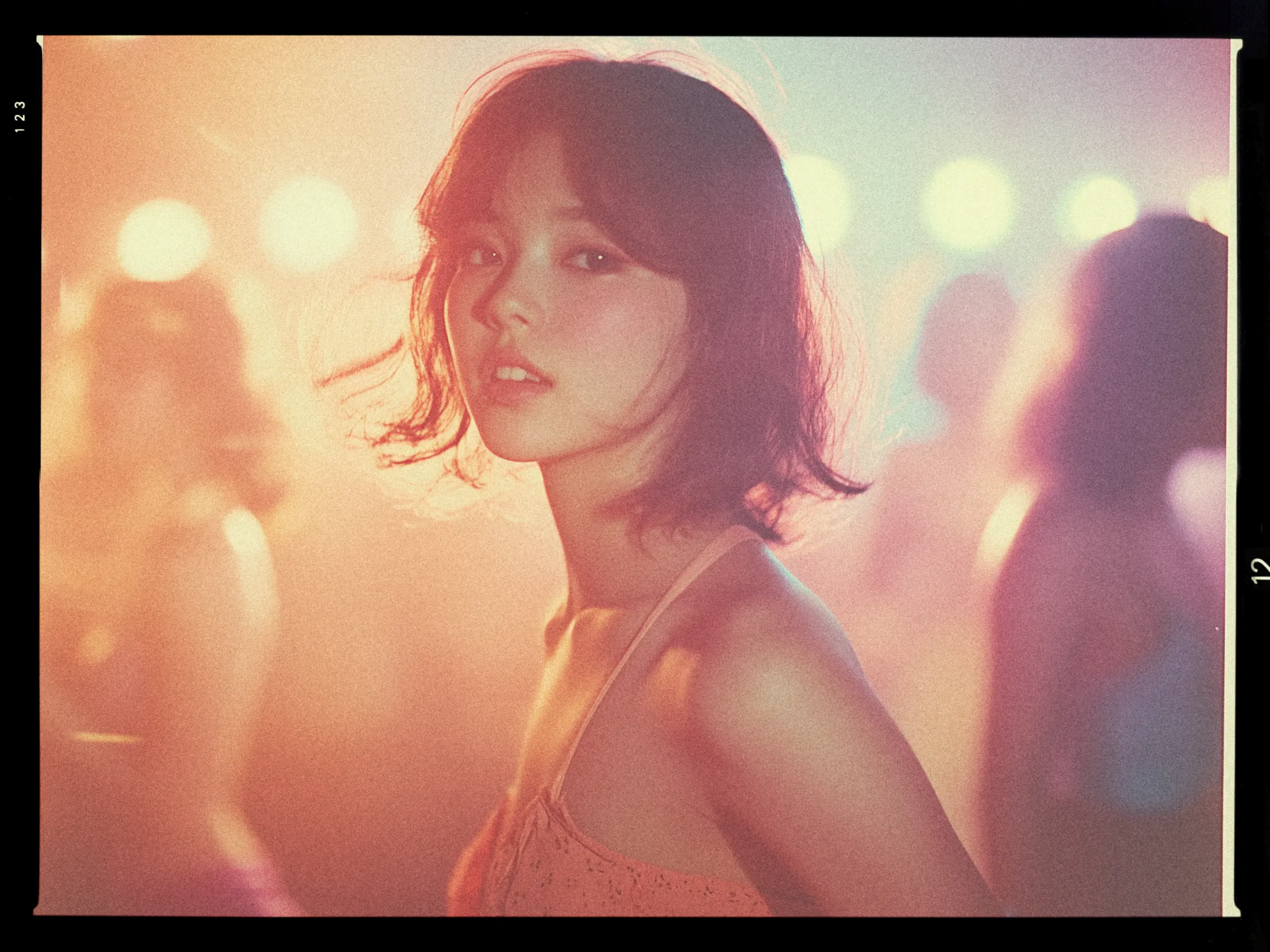 A young woman with short, wavy hair is looking towards the camera in a softly lit environment. The image has a warm, ethereal glow with a blend of pink, orange, and purple hues. She is wearing a sleeveless top or dress with thin straps. The background is softly blurred with circular lights, suggesting a lively, possibly social setting. Her expression is calm and slightly inquisitive. The overall composition has a nostalgic, dreamy quality.