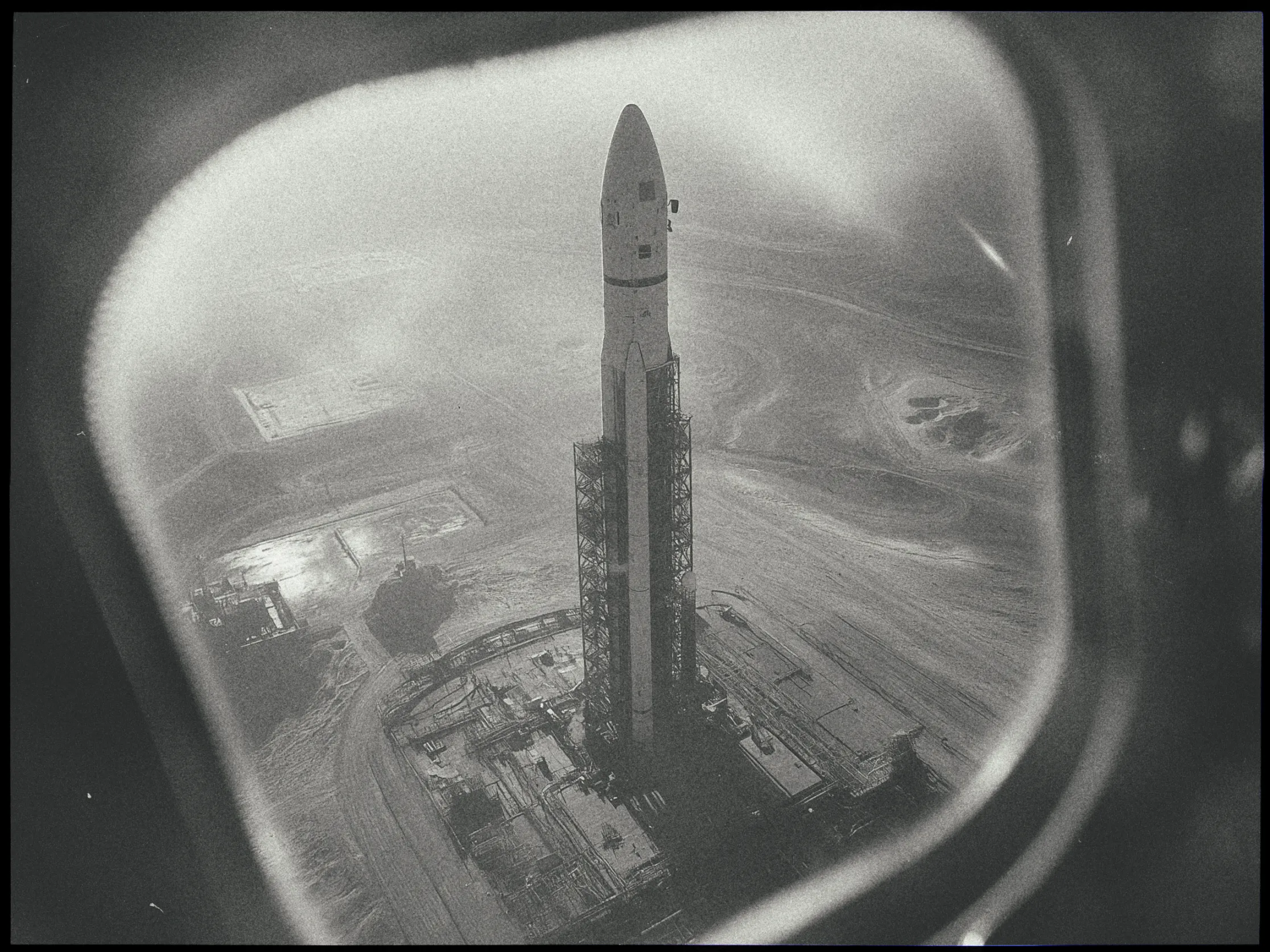 A black and white photograph showcasing a rocket on a launchpad, viewed from a high angle through a window. The rocket is upright and centered in the frame, surrounded by scaffolding and launch structures. The background shows an expansive, desolate landscape with hints of construction and roads. The window through which the image is taken adds a soft, blurred frame around the scene, emphasizing the height and distance from which the photo was captured. The overall atmosphere is historic and industrial.