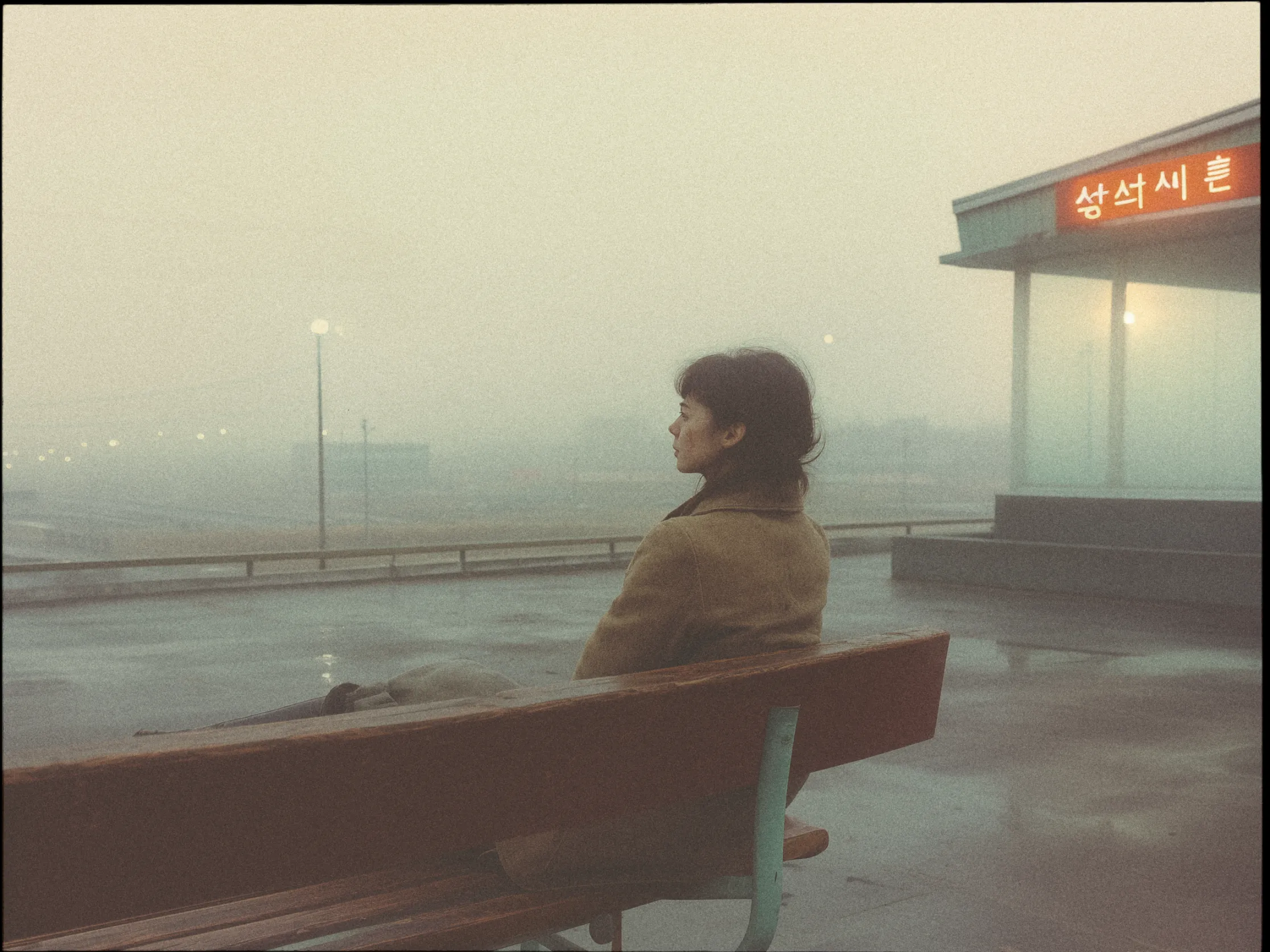 A person sits alone on a bench at a bus stop, gazing into the foggy distance. The scene has a muted, vintage tone, enhancing the sense of solitude and calm. The person is wearing a long coat, and their short hair is gently tousled by the breeze. The bus stop structure has a roof with an illuminated sign featuring Korean characters. Soft streetlights are visible through the mist, creating an atmospheric and introspective mood. The ground is wet, suggesting recent rain, and the overall ambiance is serene and contemplative.