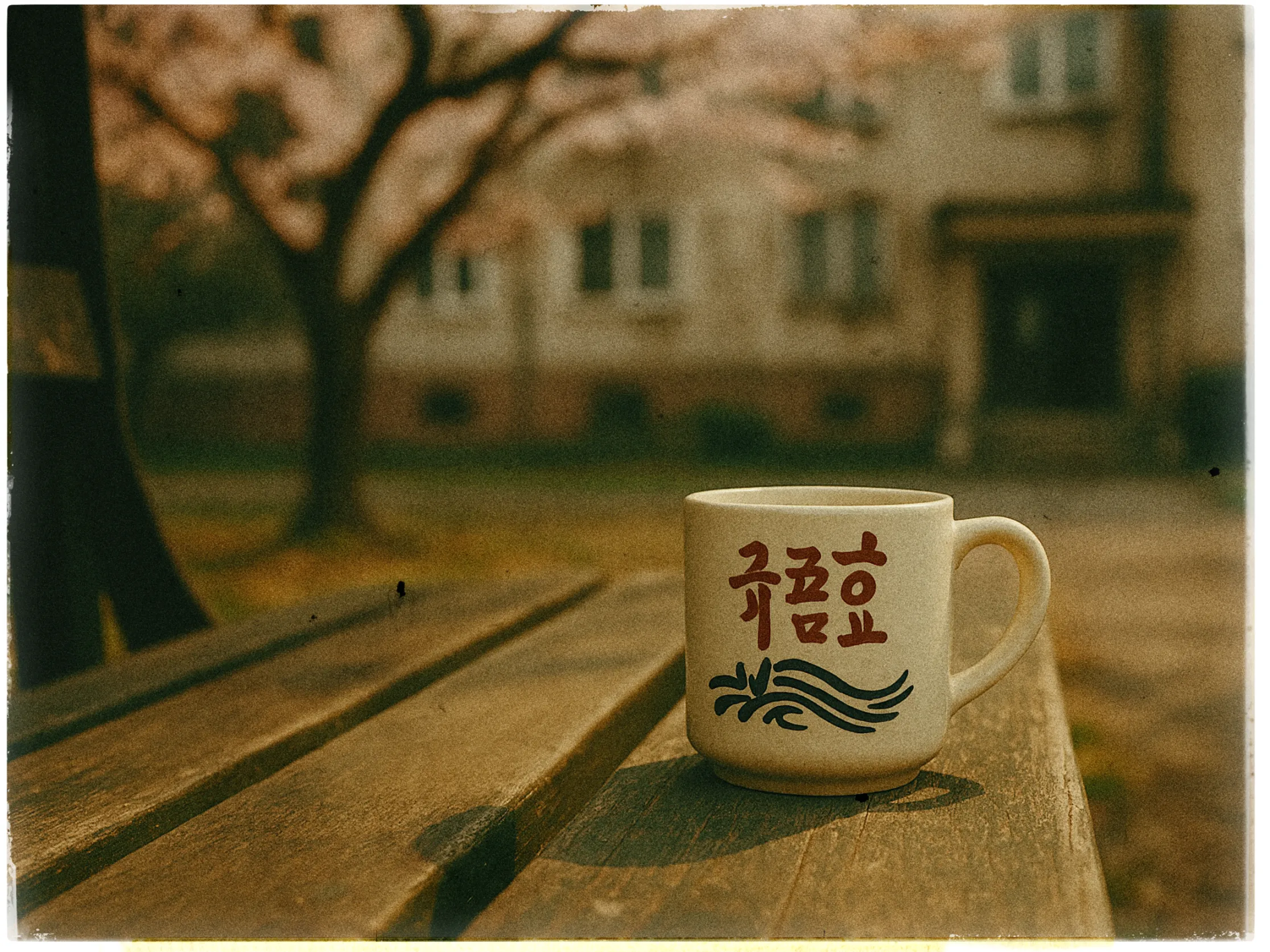 A ceramic mug with red and black designs sits on a wooden bench, with an outdoor setting in the background. The mug features stylized text and a decorative wave pattern. Out of focus, cherry blossom trees are visible, alongside a building with multiple windows and a doorway, creating a serene, nostalgic atmosphere.